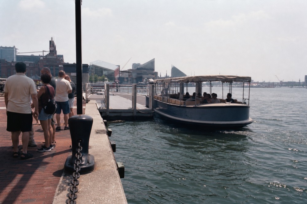 A hop-on hop-off harbor launch pulling alongside a floating dock at Harborplace, a few riders waiting on the boardwalk under a bright summer sky