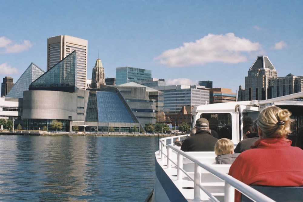 A narrated white motor cruise boat passing the National Aquarium glass pavilion, passengers at the rail, calm Inner Harbor water and a bright midday sky