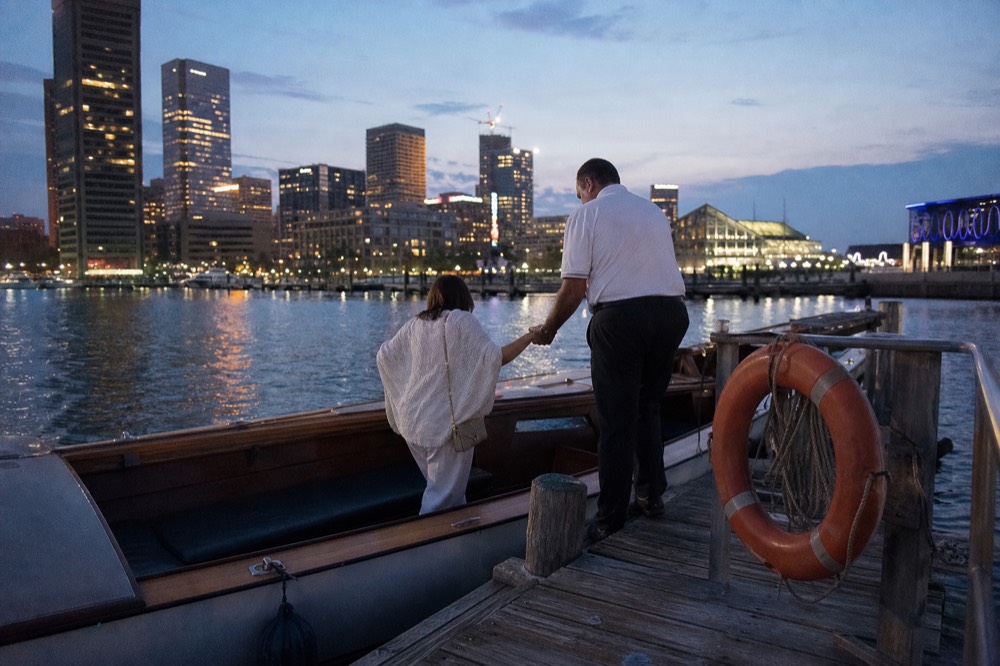 A licensed crew member in a plain polo helping a rider step aboard a low open launch from a wooden pier, an orange life ring on the rail, late afternoon light