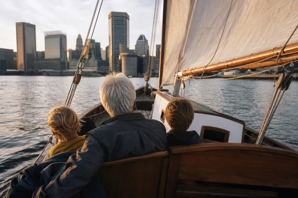 A grandparent and two children on the cockpit bench of a small schooner sailing gently across calm harbor water, the downtown skyline in soft afternoon light