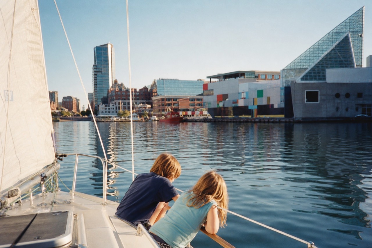 Children leaning over the rail of a small sailboat watching the water on a sunny afternoon in the Inner Harbor, the National Aquarium pavilions bright behind them