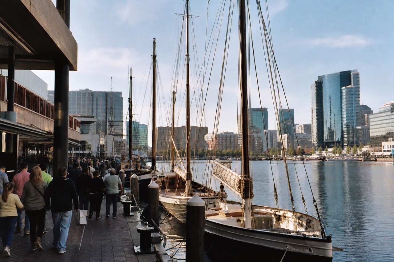 The Inner Harbor promenade busy with visitors on a bright afternoon, moored historic sailing ships and glass towers framing the open waterfront basin