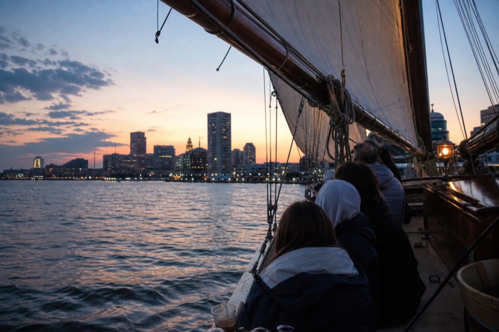 A small group seated along the rail of a schooner heeling gently under full sail, late-afternoon sun low over the Patapsco River