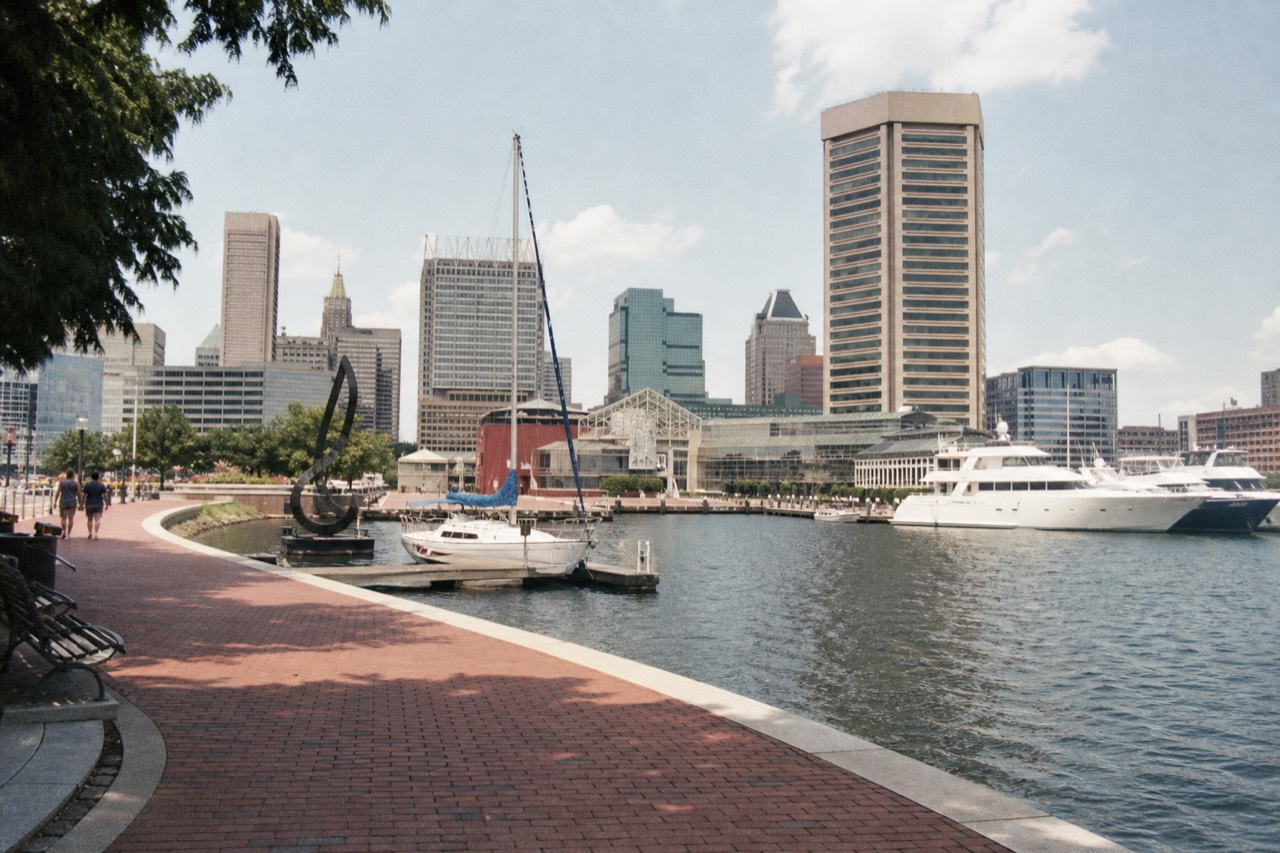 The 2.5-mile brick promenade curving around the Inner Harbor at midday, benches and public art along the water, moored boats and the downtown skyline behind