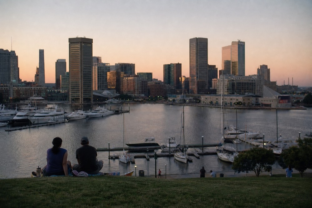 Federal Hill Park's grassy overlook above the Inner Harbor, the full downtown skyline and moored boats spread below in warm afternoon light