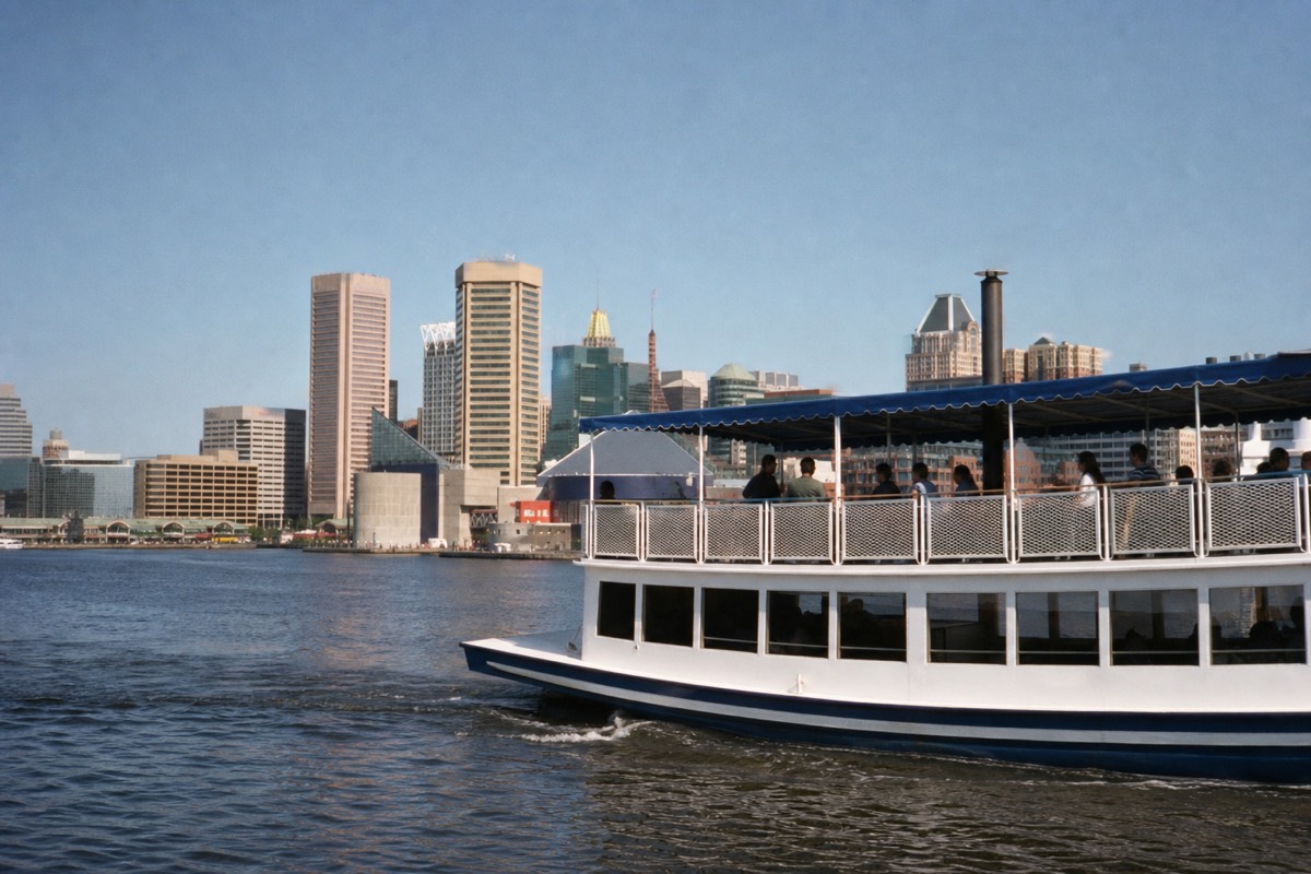 A covered sightseeing cruise boat on the Inner Harbor passing the downtown waterfront, passengers on the open upper deck under a clear sky