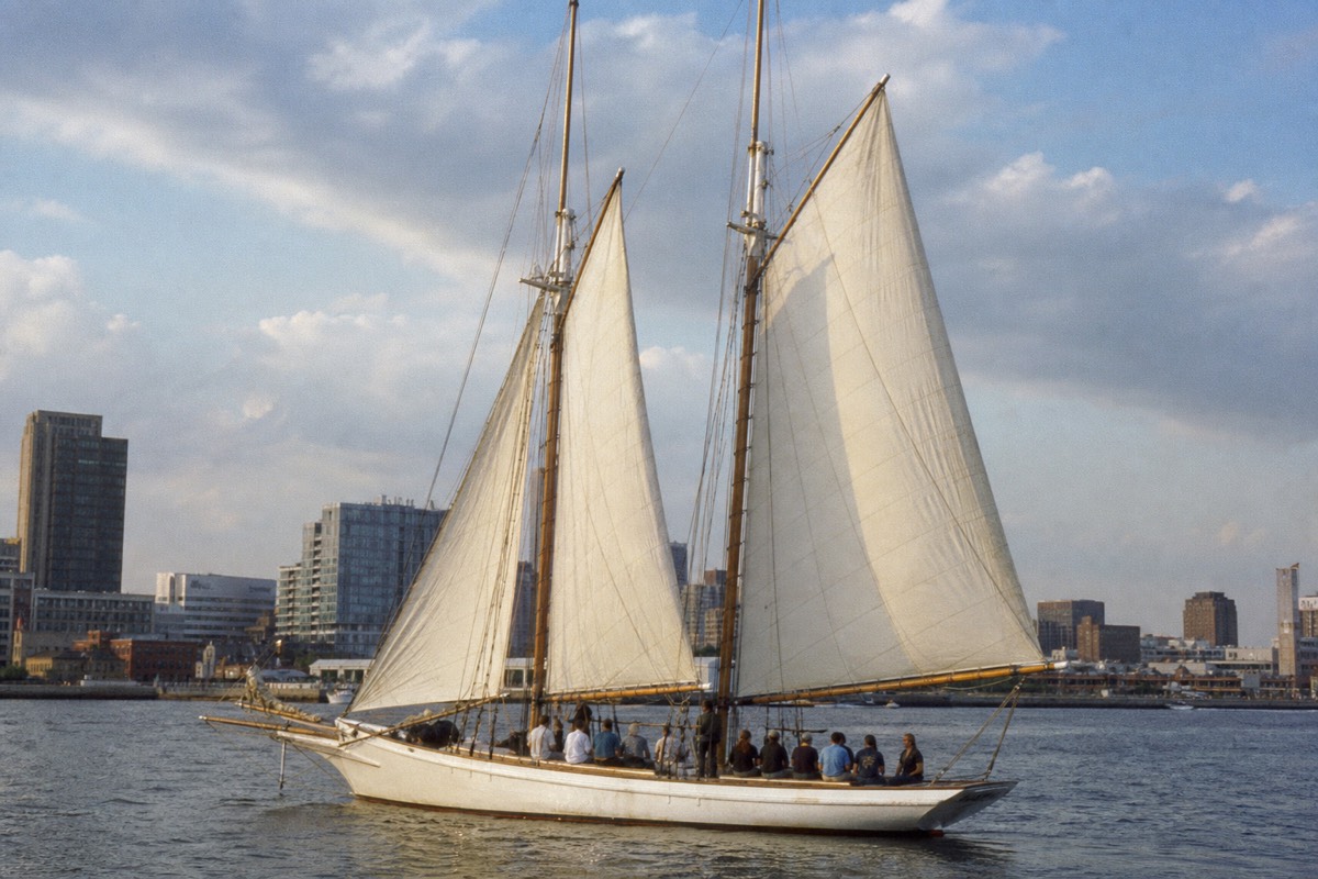 A two-masted wooden schooner under full sail on Baltimore harbor water, a small group of guests aboard, the harbor skyline low behind the rigging