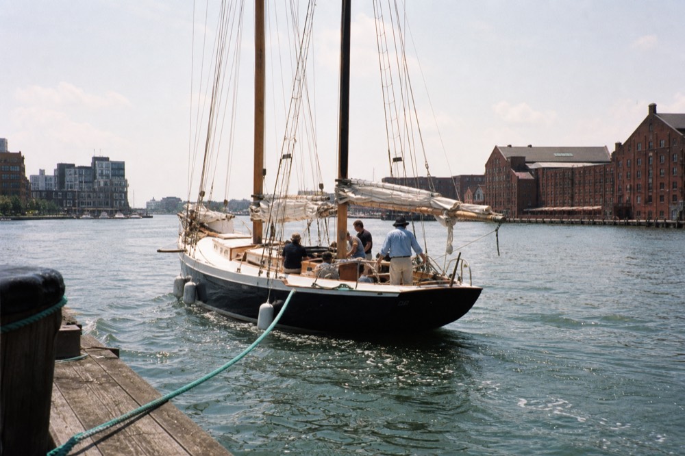 A crewed schooner casting off from a marina dock with a small group settling into the cockpit, dock lines trailing in green water, brick warehouses beyond