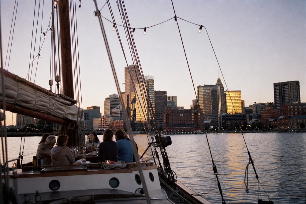 A private schooner anchored for the evening with a group's picnic spread on the cockpit table, string lights strung in the rigging, dusk over the harbor