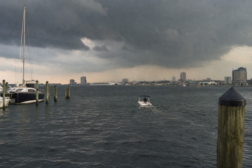 A summer thunderstorm massing in dark clouds over the Patapsco River, choppy grey water and a single small boat heading back toward the harbor mouth