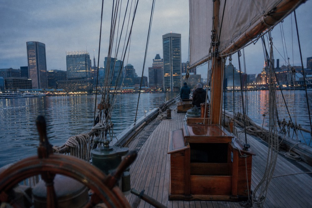 The open teak deck of a traditional sailing schooner at dusk, coiled lines and a spoked wooden wheel in the foreground, city lights beginning to reflect on dark water