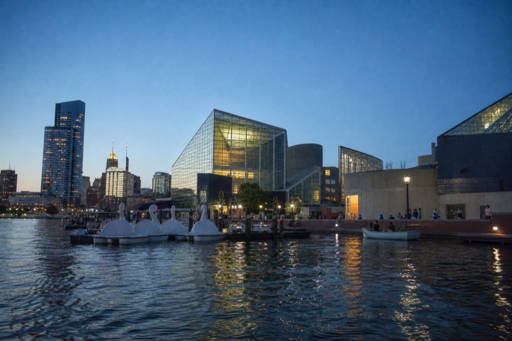 The National Aquarium's glass-and-steel pavilions seen from the water, paddleboats and a brick promenade along the waterfront, a bright open sky above