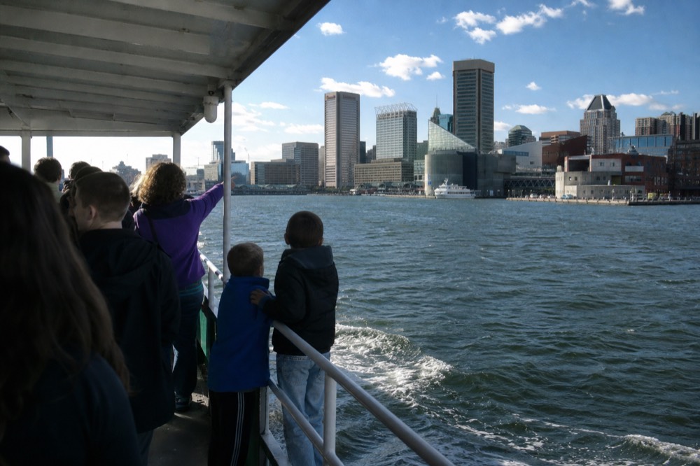 Passengers at the open rail of a covered cruise boat pointing toward the city skyline, two children on tiptoe, a breezy sunny afternoon on the harbor