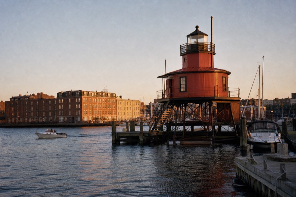 A squat red screw-pile lighthouse standing on a harbor pier with small boats tied nearby, calm water and a clear sky, a historic light preserved as a museum