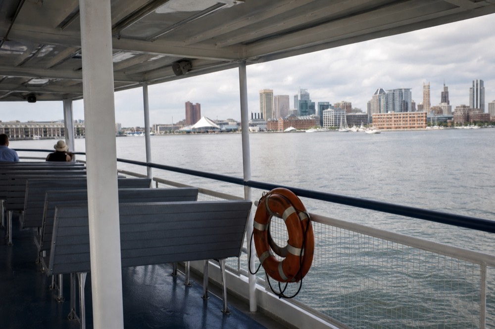 The open upper deck of a covered cruise boat with rows of empty bench seats and a life ring on the rail, the calm harbor and skyline beyond under a bright midday sky