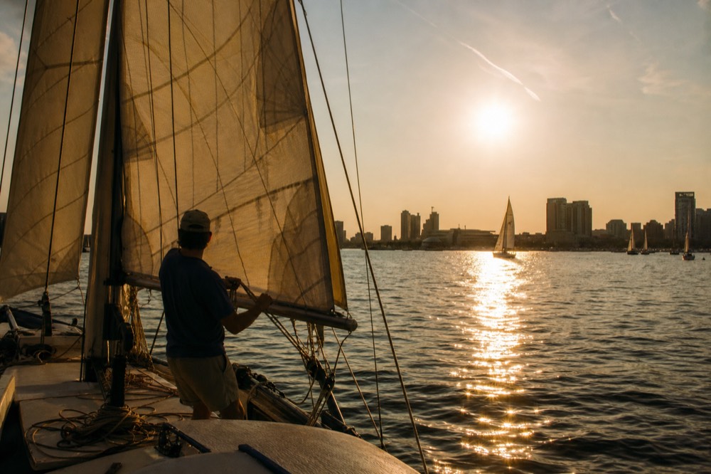 A skipper at the wheel of a small schooner trimming a line as the sun lowers, warm rim light on the sails, the harbor water turning copper