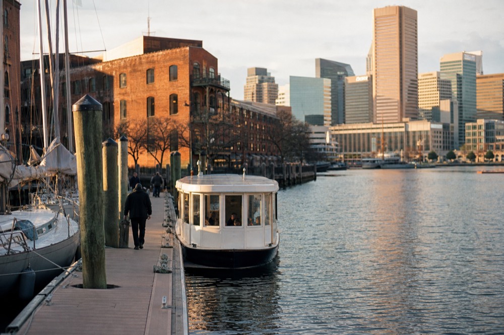 A floating dock at Fells Point with a small passenger launch idling alongside, cobblestone street and brick warehouses behind, soft morning light on the water