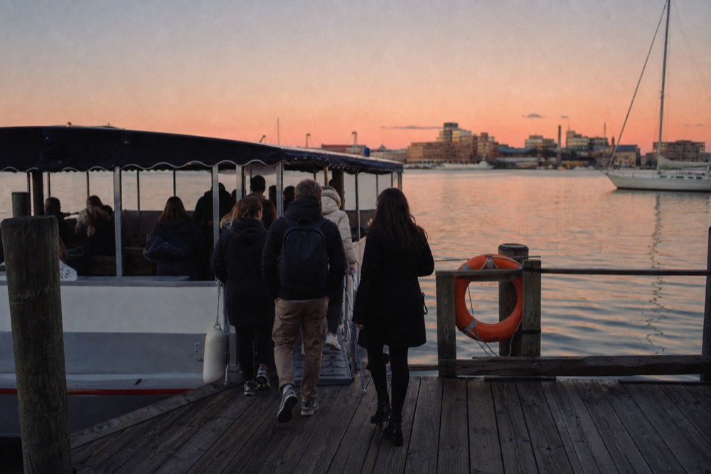 Passengers stepping onto a low open-sided harbor shuttle from a wooden pier, an orange life ring on the rail, flat calm water in the morning