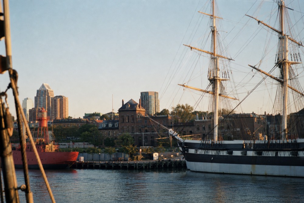 The masts and rigging of a historic naval sloop-of-war seen from a passing boat, brick piers and a red lightship behind it, gulls wheeling over the calm harbor