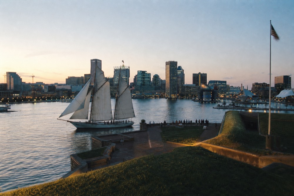 A schooner sailing close past the star-shaped earthworks of a historic waterfront fort, the American flag on a tall pole and low afternoon sun raking across the grass ramparts