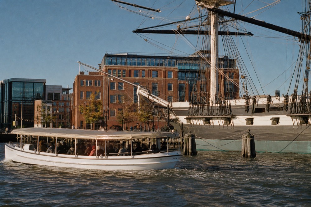 A covered white sightseeing boat passing the tall masts of a historic sloop-of-war at a downtown pier, brick waterfront buildings rising behind under a bright sky