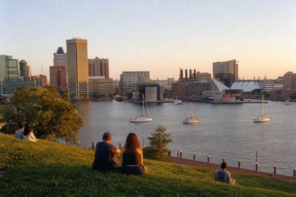 A grassy park bluff above the harbor at golden hour, a few people sitting on the slope looking out at the skyline and the water turning warm below