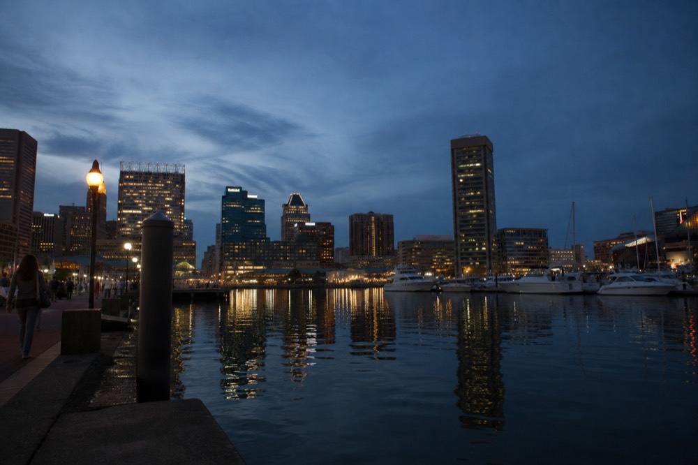 Blue-hour light over the Inner Harbor, office towers glowing gold against a deep blue sky and their reflections stretching across glassy dark water